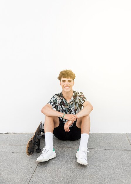 Portret van lachende man zit met skateboard tegen witte muur