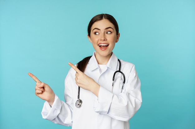 Portret van lachende medische werker, meisje arts in witte jas met stethoscoop, wijzende vingers naar links, medische kliniek advertentie tonen, torquoise achtergrond