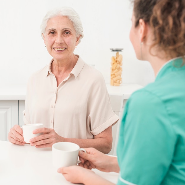 Portret van lachende senior vrouw zitten met verpleegster die koffiekopje