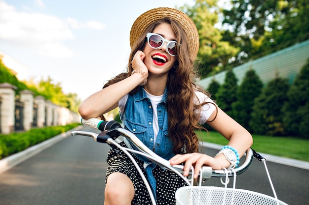 Portret van leuk meisje met lang krullend haar in hoed die een fiets op weg drijft. Ze draagt een lange rok, jerkin, blauwe zonnebril. Ze lacht naar de camera.