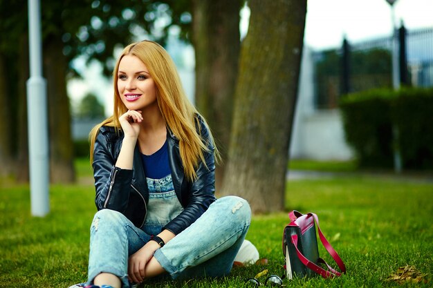 portret van leuke grappige blonde moderne sexy stedelijke jonge stijlvolle lachende vrouw meisje model in heldere moderne doek buiten zitten in het park in spijkerbroek met roze tas