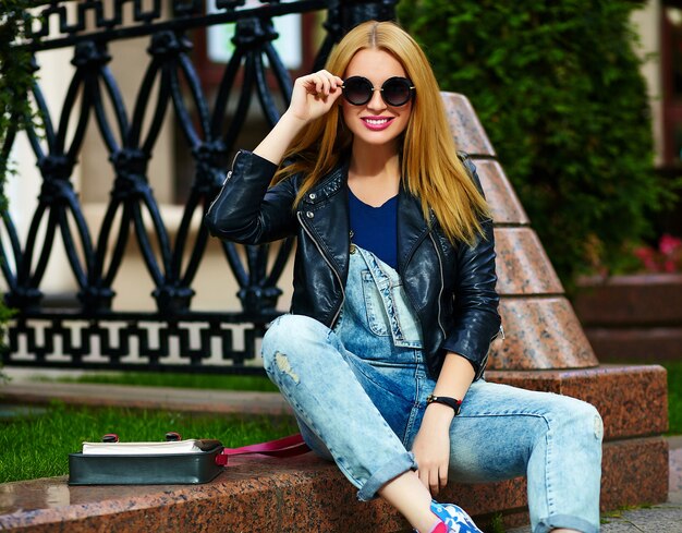 Portret van leuke grappige moderne sexy stedelijke jonge stijlvolle lachende vrouw meisje model in heldere moderne doek buiten zitten in het park in jeans op een bankje in glazen