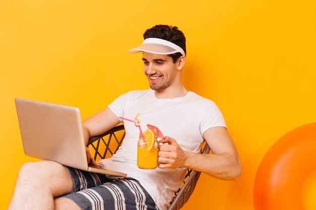 Portret van man in wit t-shirt die in laptop werkt terwijl hij op vakantie is. guy drinkt oranje cocktail.