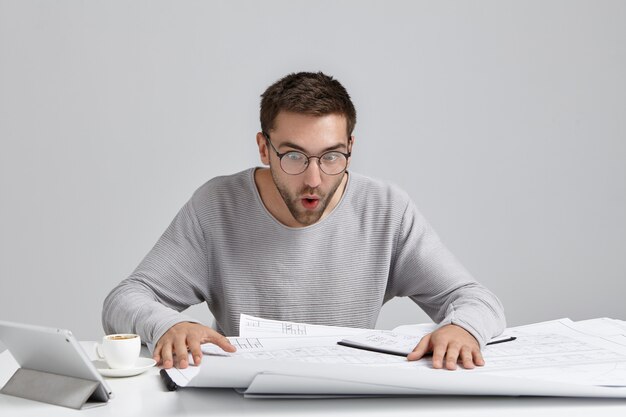 Portret van mannelijke ingenieur staart naar tekeningen, kijkt verbaasd, probeert te begrijpen wat er staat