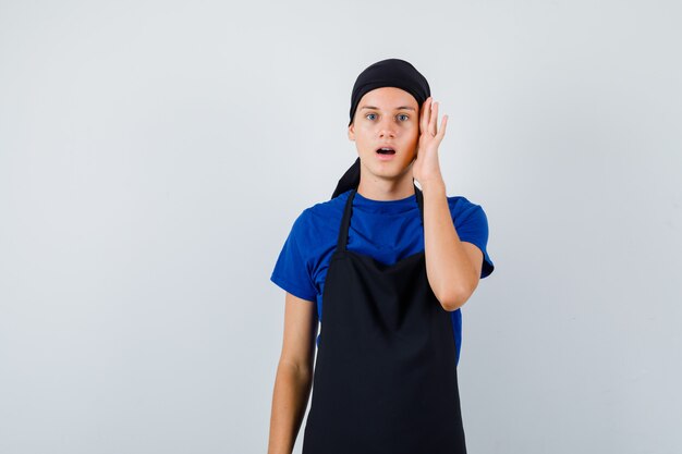 Portret van mannelijke tienerkok met hand op hoofd, mond openend in t-shirt, schort en geschokt vooraanzicht