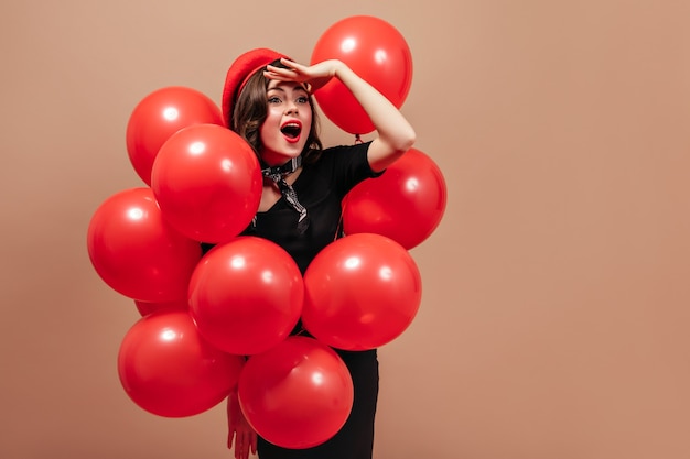 Portret van meisje met rode lippen poseren met ballonnen. Vrouw in baret onderzoekt afstand.