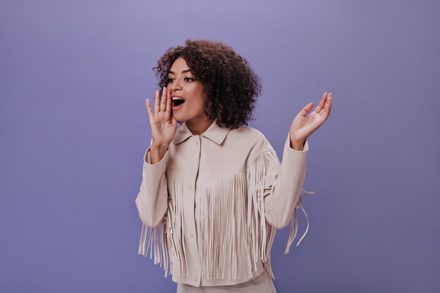 Portret van meisje schreeuwen op paarse achtergrond Aantrekkelijke jonge vrouw in beige jas schreeuwen en glimlachen op geïsoleerde achtergrond