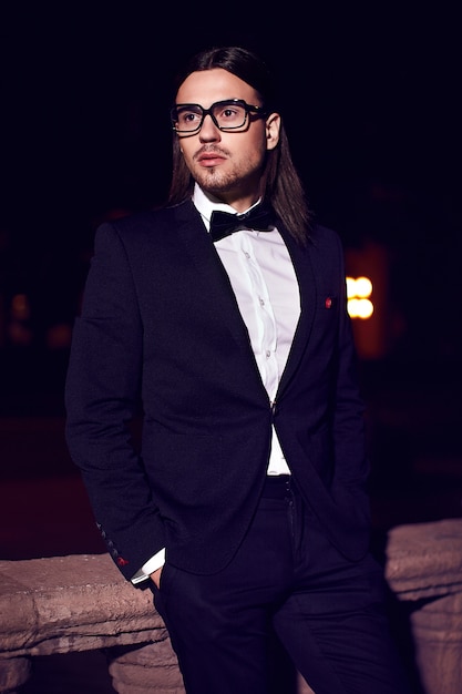 Portret van mode elegante langharige jonge man. Aantrekkelijk en knap mannelijk model in zwart pak met snor in de straat 's nachts