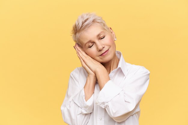 Portret van modieuze blonde vrouw met pensioen poseren geïsoleerd buigend hoofd, handpalmen onder de wang vasthouden en ogen gesloten houden, slapen, dutten, glimlachen van plezier, goede droom hebben