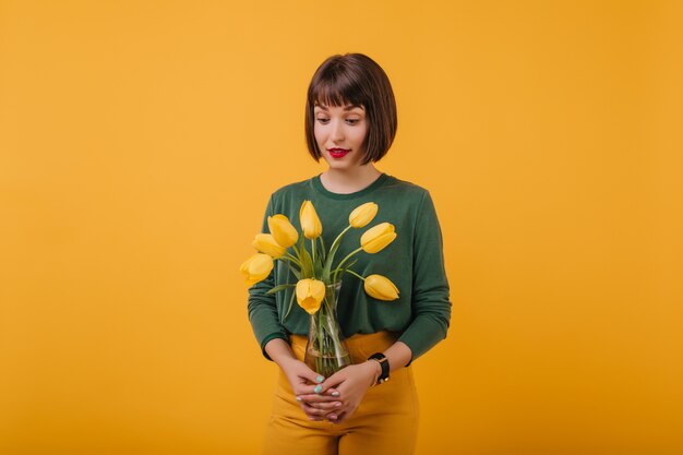 portret van modieuze dame met trendy kapsel met vaas met tulpen. Binnenfoto van geïnteresseerd donkerbruin meisje dat met bloemboeket wordt geïsoleerd.