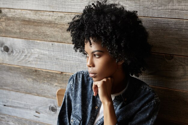 Portret van modieuze jonge Afro-Amerikaanse vrouw gekleed in denim shirt met serieuze en doordachte blik, na te denken over iets belangrijks, zittend in café met hoofd achterover op haar hand