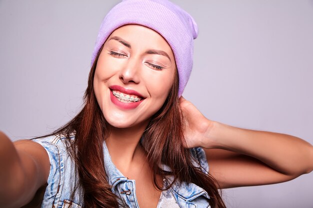 Portret van mooi glimlachend leuk donkerbruin vrouwenmodel in de toevallige kleren van de zomerjeans zonder make-up in purpere beanie die selfie foto op telefoon maken die op grijs wordt geïsoleerd