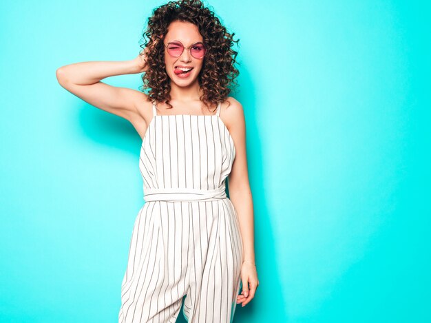 Portret van mooi glimlachend model met afro krullen kapsel gekleed in zomer hipster kleding. Sexy zorgeloos meisje poseren in de buurt van blauwe muur. Trendy grappige en positieve vrouw