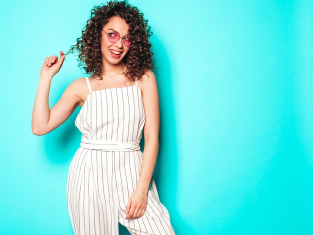 Portret van mooi glimlachend model met afro krullen kapsel gekleed in zomer hipster kleding. Sexy zorgeloos meisje poseren in de buurt van blauwe muur. Trendy grappige en positieve vrouw