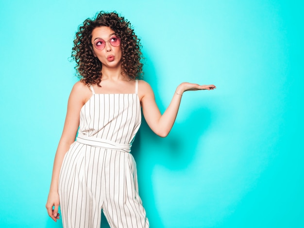 Portret van mooi glimlachend model met afro krullen kapsel gekleed in zomer hipster kleding. Sexy zorgeloos meisje poseren in de buurt van blauwe muur. Trendy vrouw iets op haar hand demonstreren