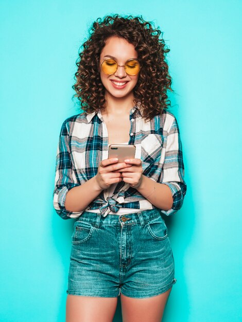 Portret van mooi glimlachend model met afro krullen kapsel gekleed in zomerkleren. Zorgeloos meisje poseren in de buurt van blauwe muur. Vrouw gebruikt haar mobiele telefoon en sms typen. Ze zoekt naar winkelverkoop