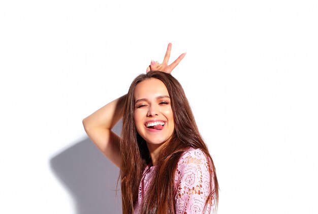 Portret van mooi Kaukasisch het glimlachen donkerbruin vrouwenmodel in heldere roze de zomer modieuze die kleding op witte achtergrond wordt geïsoleerd. Haar tong en vredesteken tonen