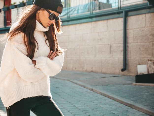 Portret van mooi lachend model. Vrouw gekleed in warme hipster witte trui en muts. Trendy vrouw die op straat poseert