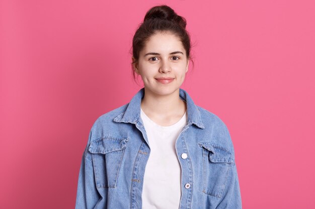 Portret van mooi meisje met broodje in spijkerjasje en wit t-shirt met zachte glimlach op roze