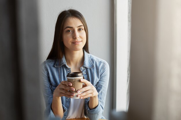 Portret van mooi ontspannen meisje met donker haar in witte t-shirt onder denimoverhemd die terwijl het drinken van kop van koffie glimlachen.