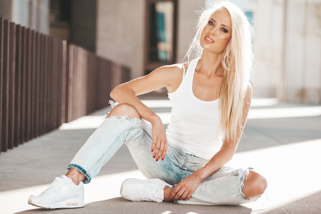 Portret van mooi schattig blond meisje in witte T-shirt en jeans die in openlucht stellen. Leuke meisjeszitting op asfalt op de straat