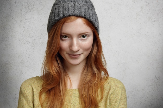 Portret van mooi sproeterig meisje met lang rood haar en schattige charmante glimlach met grijze stijlvolle hoed kijken en glimlachend staande tegen muur