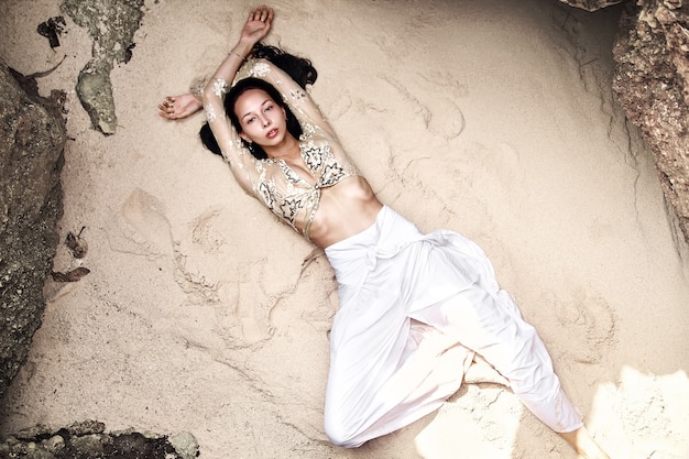 portret van mooie blanke vrouw model met donker lang haar in klassieke wijde pijpen broek liggend op wit zand op het strand in de buurt van rotsen. Bovenaanzicht