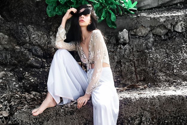 portret van mooie blanke vrouw model met lang donker haar in wijde pijpen klassieke broek poseren in de buurt van rotsen en groene tropische exotische bladeren achtergrond