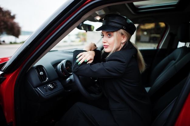 Portret van mooie blonde sexy mode vrouw model in pet en in helemaal zwart met lichte make-up zitten en rijden rode stadsauto