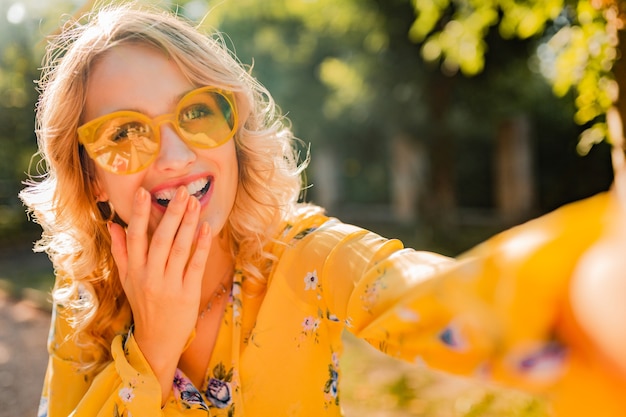 Gratis foto portret van mooie blonde stijlvolle lachende vrouw in gele blouse dragen van een zonnebril selfie foto maken