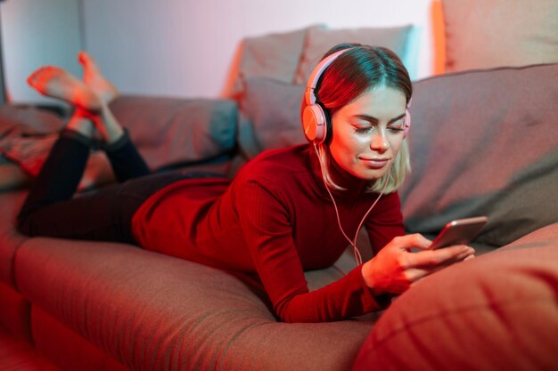 Portret van mooie dame liggend op de bank in koptelefoon en mobiel in handen Jonge coole vrouw die muziek luistert terwijl ze thuis tijd doorbrengt