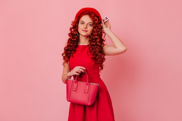 Portret van mooie dame met golvend rood haar en blauwe ogen. Meisje in een rode jurk zet haar baret op en houdt haar tas vast.
