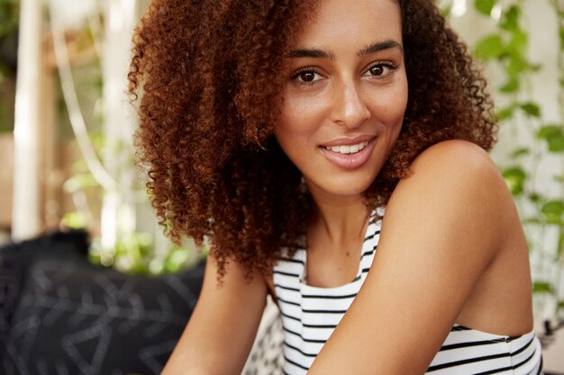 Portret van mooie donkere vrouw met Afro kapsel close-up heeft zelfverzekerde vrolijke uitstraling, besteedt vrije tijd in cafetaria. Goed uitziende jonge Afro-Amerikaanse vrouw recreëren na het werk