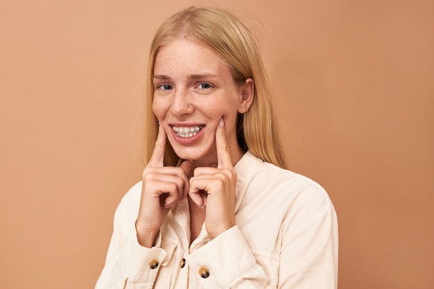 Portret van mooie gefrustreerde jonge vrouw met lang blond haar en neuspiercing met pijnlijke gezichtsuitdrukking terwijl haar tandvlees pijn doet vanwege strakke tandenbeugels