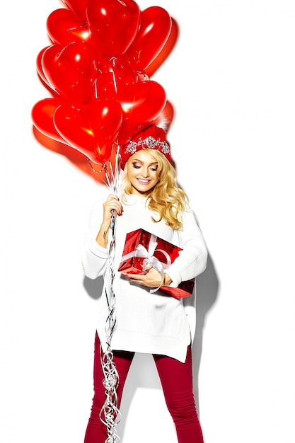 Portret van mooie gelukkig lieve lachende blonde vrouw meisje houdt in haar handen grote kerstcadeau doos en hart ballonnen in casual rode hipster winterkleren, in witte warme trui