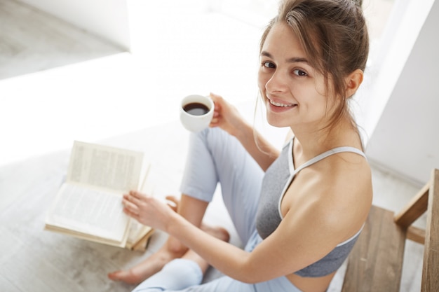 Portret van mooie gelukkige vrouw die lacht houden kopje koffie zittend op de vloer met boek.