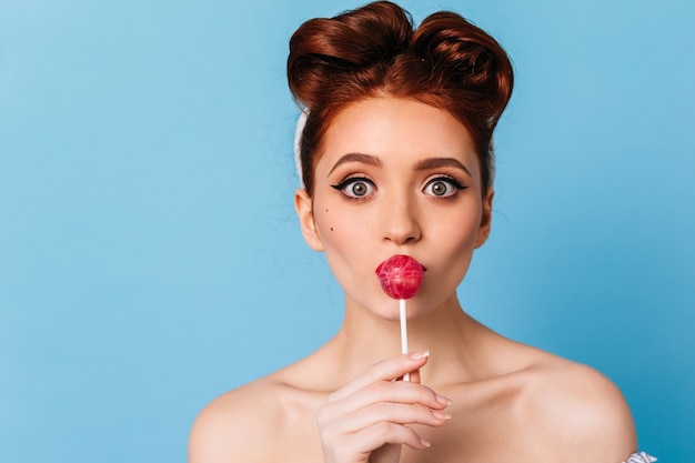 Gratis foto portret van mooie gember jonge vrouw die lolly likken. charmant kaukasisch meisje dat snoep eet op blauwe ruimte.