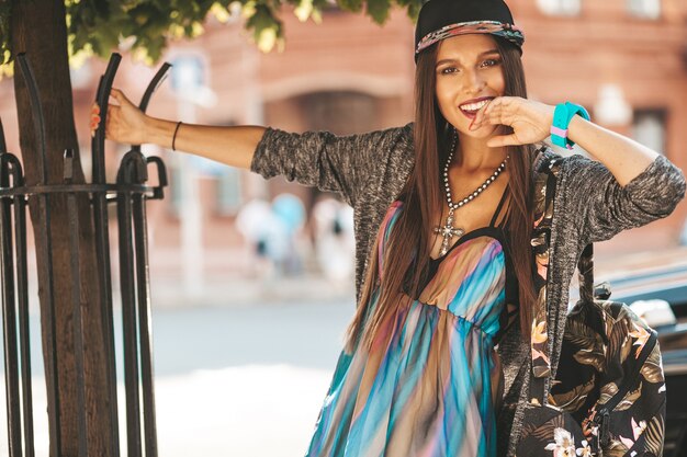 Portret van mooie glamour lachende brunette tiener model in zomer hipster kleding en tas. Meisje poseren in de straat. Vrouw in cap