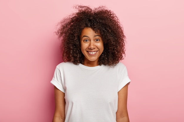 Portret van mooie jonge Afro-Amerikaanse vrouw glimlacht positief, geniet van grappige scène, wordt geamuseerd, grinnikt over hilarische grap, heeft donker krullend volumineus haar, draagt casual outfit