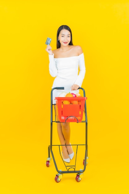 Portret van mooie jonge Aziatische bedrijfsvrouw met winkelmandje met kruidenierswinkel van supermarkt op gele muur