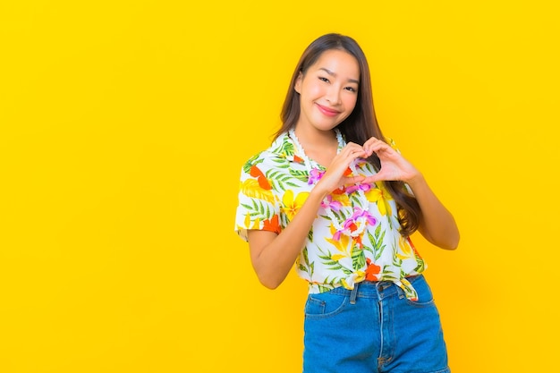 Portret van mooie jonge Aziatische vrouw die kleurrijk overhemd draagt en hart op gele muur maakt ondertekenen