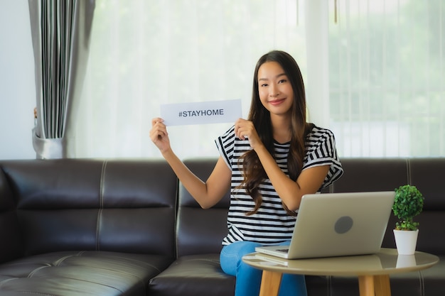 Portret van mooie jonge Aziatische vrouw die laptop op bank met het huisdocument van het verblijf met behulp van