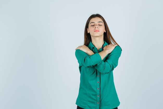 Portret van mooie jonge dame die handen op schouders houdt, ogen in groen overhemd sluit en ontspannen vooraanzicht kijkt