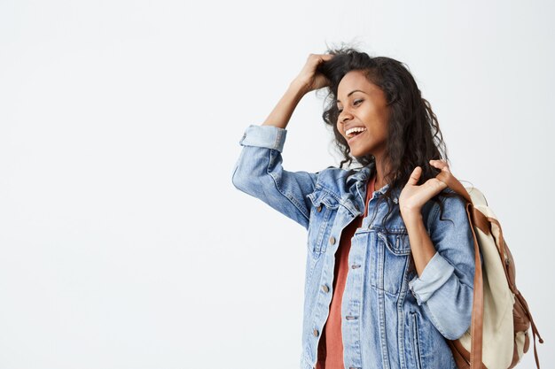 Portret van mooie jonge donkere vrouw met golvend donker haar, gekleed in casual rood T-shirt en spijkerjasje met rugzak op haar schouder aangenaam lachend. Trendy hipster vrouw met goed humeur