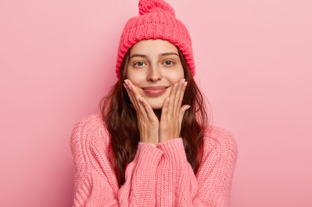 Portret van mooie jonge vrouw glimlacht aangenaam, houdt beide handpalmen op de wangen, kijkt graag naar de camera, heeft een ontspannen blik, draagt gebreide muts en trui, modellen binnen over roze studiomuur