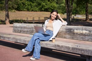 Portret van mooie jonge vrouw zittend op een bankje in het park met behulp van laptop en koffie drinken ontspannen