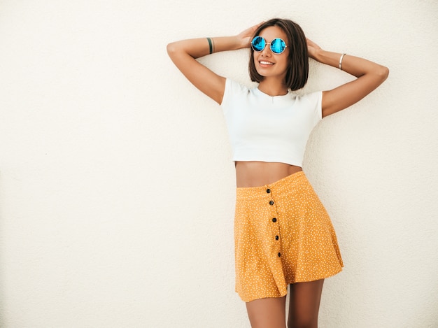 Gratis foto portret van mooie lachende brunette model gekleed in zomer hipster kleding. trendy meisje poseren in de straat in de buurt van witte muur. grappige en positieve vrouw die pret heeft