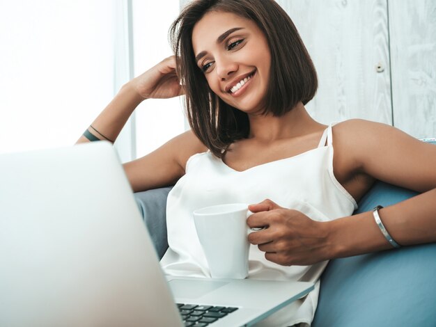 Portret van mooie lachende vrouw gekleed in witte pyjama. Zorgeloos model zittend op een zachte tas stoel en met behulp van laptop.