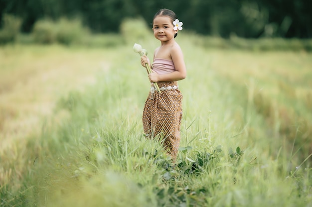 Portret van mooie meisjes in Thaise traditionele kleding en witte bloem op haar oor gezet, staande en twee lotus in de hand houdend op rijstveld, ze glimlacht van geluk, kopieer ruimte