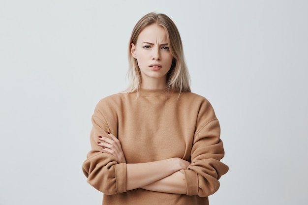 portret van mooie vrouw met blond steil haar fronst haar gezicht in ongenoegen, draagt een losse trui met lange mouwen en houdt de armen over elkaar. Aantrekkelijke jonge vrouw in gesloten houding.